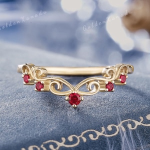 May include: A gold-colored ring with a curved design, featuring small red gemstones. The ring has an ornate, filigree-style pattern with a central gemstone and additional stones along the curve. The text "GOLDENXANADU" is at the top.