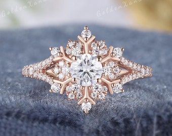 May include: A rose gold snowflake engagement ring with a round center stone and smaller stones set in the band and snowflake design.  The ring is on a gray textured surface.