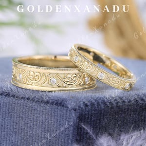 May include: Two gold wedding rings with intricate floral and scrollwork designs, each set with small diamonds. The rings are displayed on a blue velvet surface. The text "GOLDENXANADU" is at the top.