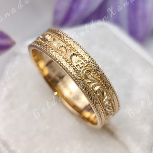 Pode incluir: Um anel de ouro com intrincados padrões de volutas gravados ao longo da banda externa, contornado por detalhes finos de milgrain. O texto GOLDENXANADU é visível no topo da imagem e o anel repousa sobre uma caixa de veludo branca.