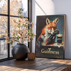 May include: A framed vintage Guinness advertisement featuring a fox with a pint of stout. The artwork is displayed on a wooden surface, next to a black vase with floral branches and a small wooden cup. The scene is illuminated by natural light from a nearby window.