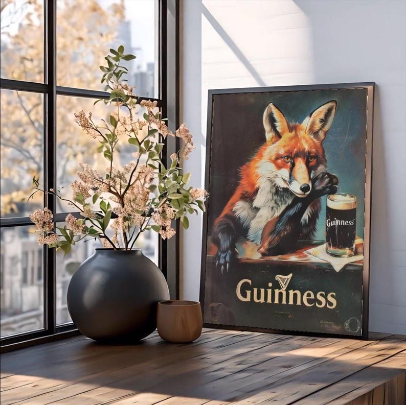 May include: A framed vintage Guinness advertisement featuring a fox with a pint of stout. The artwork is displayed on a wooden surface, next to a black vase with floral branches and a small wooden cup. The scene is illuminated by natural light from a nearby window.
