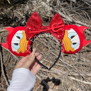 May include: Red devil ears with a red sequin bow and a red trident. The ears feature a cartoon duck face.