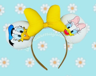 Puede incluir: Una diadema blanca con un lazo amarillo y dos orejas blancas. Una oreja presenta una imagen de dibujos animados de Donald Duck y la otra oreja presenta una imagen de dibujos animados de Daisy Duck.