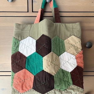 May include: A green canvas tote bag with a patchwork design of various colors, including brown, green, white, and orange. The bag has a brown corduroy handle.