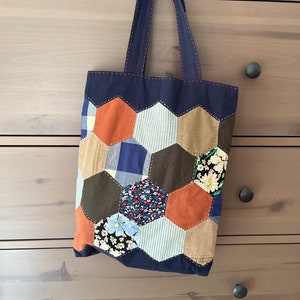 May include: A blue patchwork tote bag with a hexagonal pattern. The bag is made of different fabrics in shades of blue, brown, orange, and white. The bag has a single handle and is hanging on a wooden dresser.