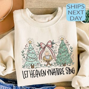 May include: Cream-colored sweatshirt featuring a Christmas-themed graphic. The design includes two green Christmas trees, a pink bow, and a depiction of baby Jesus in a manger. The text "LET HEAVEN & NATURE SING" is printed below the graphic.
