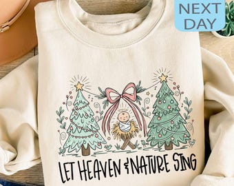 May include: A cream-coloured sweatshirt with a Christmas graphic. The design features two green Christmas trees, a pink bow, and a depiction of baby Jesus in a manger. The text "LET HEAVEN & NATURE SING" is printed below the graphic.