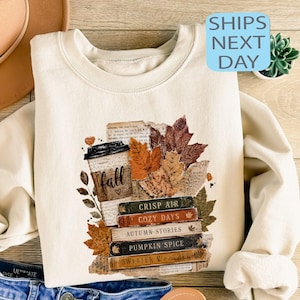 May include: A cream-colored crewneck sweatshirt featuring a graphic print of a coffee cup, autumn leaves, and a stack of books with the text: fall, crisp air, cozy days, autumn stories, pumpkin spice, and sweater weather printed on the book spines.