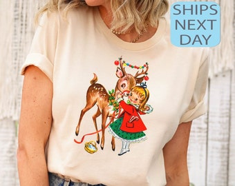 Op de afbeelding: Een wit T-shirt met een vintage illustratie van een meisje dat een rendier vasthoudt met een rode strik en een kerstlichtjeskrans. Het meisje draagt een rode jurk met een groene rok en witte schoenen.