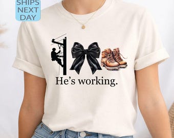 May include: A cream-coloured t-shirt with a graphic of a lineman on a utility pole, a black bow, and work boots. The text "He's working" is printed below. The shirt has short sleeves and is made of a soft material.