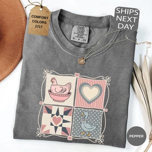 May include: A grey Comfort Colors t-shirt with a patchwork design featuring a chicken in a bowl, a heart, and a duck. The shirt has a crew neck and short sleeves. A tag reads "Comfort Colors 1717". A necklace is draped across the shirt.