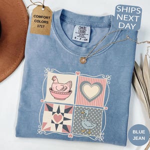 May include: Blue jean coloured t-shirt with a patchwork design featuring a hen, heart, and duck. The shirt has a Comfort Colors tag and a small tag that says "SHIPS NEXT DAY". A gold necklace is draped across the shirt.