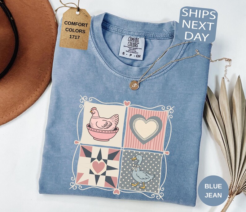 May include: Blue jean coloured t-shirt with a patchwork design featuring a hen, heart, and duck. The shirt has a Comfort Colors tag and a small tag that says "SHIPS NEXT DAY". A gold necklace is draped across the shirt.
