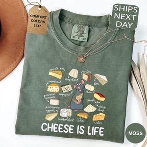 May include: A moss green t-shirt with a graphic of various cheeses and a cartoon rat holding a cheese wedge. The text "CHEESE IS LIFE" is printed below the graphic. The shirt has a crew neck and short sleeves.