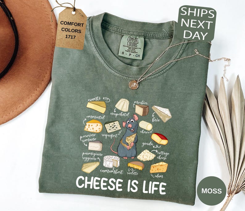 May include: A moss green t-shirt with a graphic of various cheeses and a cartoon rat holding a cheese wedge. The text "CHEESE IS LIFE" is printed below the graphic. The shirt has a crew neck and short sleeves.