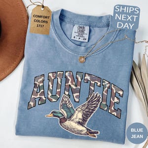 May include: A blue jean Comfort Colors t-shirt with the word "AUNTIE" in camouflage lettering and a duck graphic. The shirt has a crew neck and short sleeves. A gold necklace is draped across the shirt.