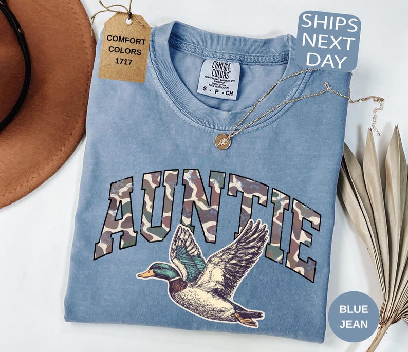 May include: A blue jean Comfort Colors t-shirt with the word "AUNTIE" in camouflage lettering and a duck graphic. The shirt has a crew neck and short sleeves. A gold necklace is draped across the shirt.