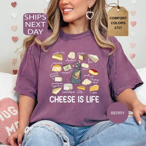May include: Berry-colored t-shirt with a cheese-themed graphic. The design features various cheese illustrations, a cartoon rat, and the text "CHEESE IS LIFE". The shirt is a comfort colors style.