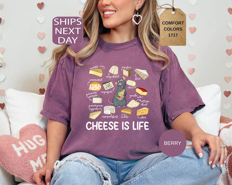 May include: Berry-colored t-shirt with a cheese-themed graphic. The design features various cheese illustrations, a cartoon rat, and the text "CHEESE IS LIFE". The shirt is a comfort colors style.