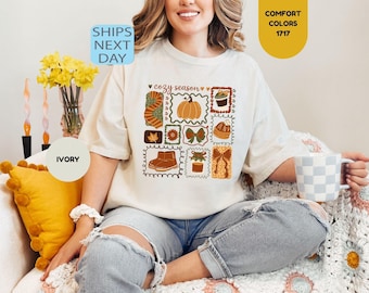 May include: An ivory t-shirt featuring a graphic print with the text cozy season. The design includes a grid of illustrations depicting a pumpkin, a scarf, a leaf, a flower, a bow, boots, a cupcake, a beanie, and a gift box in autumn colors.