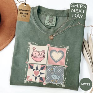 May include: A moss green Comfort Colors t-shirt with a patchwork design featuring a hen, heart, and duck. The shirt has a crew neck and short sleeves. A gold necklace is draped across the shirt. The tag reads "Comfort Colors 1717".