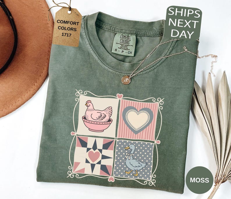 May include: A moss green Comfort Colors t-shirt with a patchwork design featuring a hen, heart, and duck. The shirt has a crew neck and short sleeves. A gold necklace is draped across the shirt. The tag reads "Comfort Colors 1717".