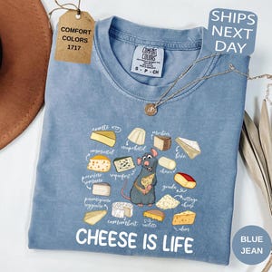 May include: A blue jean Comfort Colors t-shirt with a graphic of various cheeses and a cartoon rat holding cheese. The text "CHEESE IS LIFE" is printed below the graphic. A tag reads "COMFORT COLORS 1717".