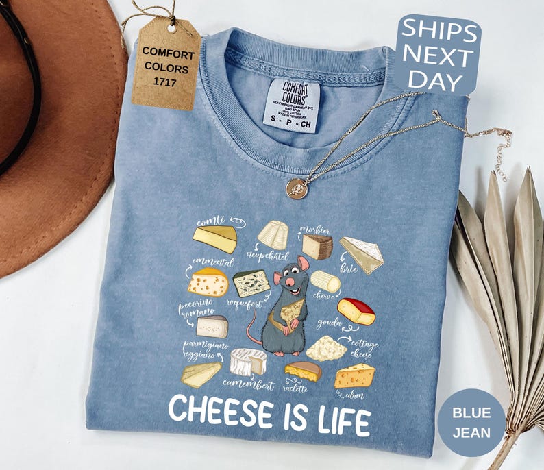 May include: A blue jean Comfort Colors t-shirt with a graphic of various cheeses and a cartoon rat holding cheese. The text "CHEESE IS LIFE" is printed below the graphic. A tag reads "COMFORT COLORS 1717".