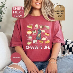 May include: Crimson-colored t-shirt featuring a cartoon illustration of various cheeses and a gray rat. The text "CHEESE IS LIFE" is printed below the cheese illustrations. The shirt is part of the Comfort Colors 1717 collection.