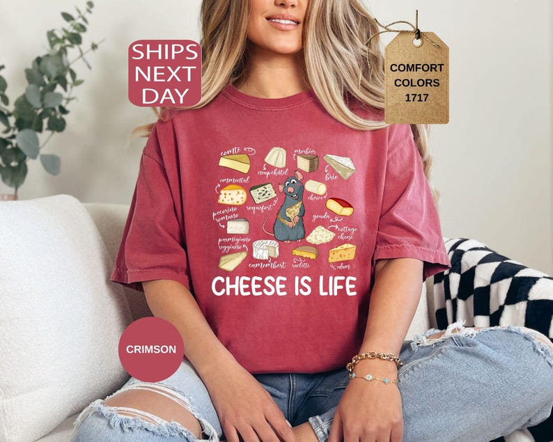 May include: Crimson-colored t-shirt featuring a cartoon illustration of various cheeses and a gray rat. The text "CHEESE IS LIFE" is printed below the cheese illustrations. The shirt is part of the Comfort Colors 1717 collection.