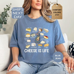 May include: Blue jean-colored t-shirt featuring a cartoon rat holding cheese, surrounded by illustrations of various cheeses. The text "CHEESE IS LIFE" is printed below the graphic. The shirt is a Comfort Colors brand.