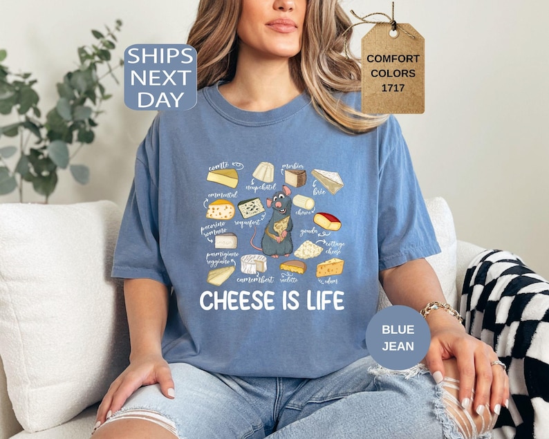 May include: Blue jean-colored t-shirt featuring a cartoon rat holding cheese, surrounded by illustrations of various cheeses. The text "CHEESE IS LIFE" is printed below the graphic. The shirt is a Comfort Colors brand.