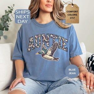 May include: Blue jean-colored t-shirt with the word "AUNTIE" in camouflage lettering and a graphic of a flying duck. The shirt has a relaxed fit and is made of a soft material. The shirt also has the words "Comfort Colors" and "1717" on a tag.