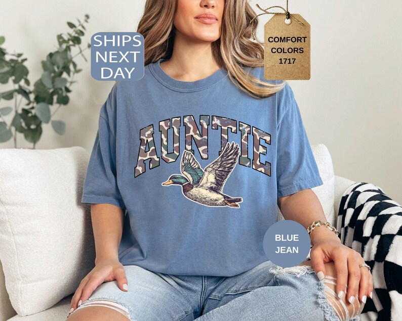 May include: Blue jean-colored t-shirt with the word "AUNTIE" in camouflage lettering and a graphic of a flying duck. The shirt has a relaxed fit and is made of a soft material. The shirt also has the words "Comfort Colors" and "1717" on a tag.