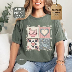 May include: A moss green t-shirt with a patchwork design featuring a hen, heart, and duck. The shirt has the text "SHIPS NEXT DAY" and a tag that says "COMFORT COLORS 1717".