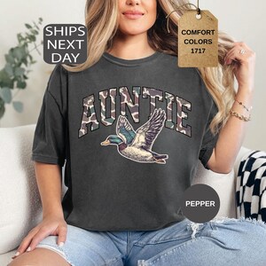 May include: Dark gray Auntie t-shirt with a camouflage design on the word "AUNTIE" and a flying duck graphic. The shirt is a comfort color and is a pepper color.