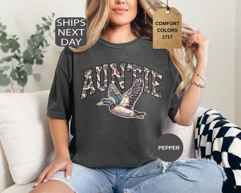 May include: Dark gray Auntie t-shirt with a camouflage design on the word "AUNTIE" and a flying duck graphic. The shirt is a comfort color and is a pepper color.