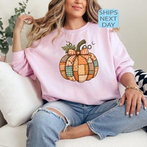 May include: A light pink sweatshirt featuring a patchwork pumpkin design with a green stem and bow. The pumpkin is made of various fabric patterns in orange, green, and brown. The text "SHIPS NEXT DAY" is in a blue box.