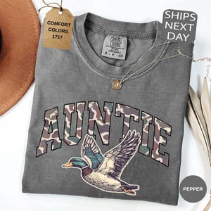 May include: A gray Comfort Colors t-shirt with the word "AUNTIE" in a camouflage design and a duck graphic. The shirt has a round neckline and short sleeves. A tag reads "Comfort Colors 1717". A necklace is draped across the shirt.