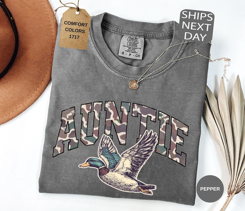 May include: A gray Comfort Colors t-shirt with the word "AUNTIE" in a camouflage design and a duck graphic. The shirt has a round neckline and short sleeves. A tag reads "Comfort Colors 1717". A necklace is draped across the shirt.