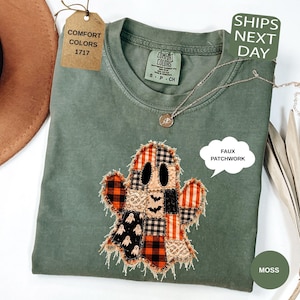 May include: A moss green short-sleeve t-shirt featuring a central graphic of a ghost constructed from various orange, black, and white patterned fabric squares. The ghost design has a frayed edge appearance and black oval eyes.