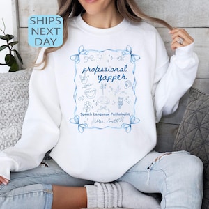 Puede incluir: Sudadera blanca con un diseño ilustrado en azul. El diseño incluye las palabras "professional yapper" y "Speech Language Pathologist" en escritura azul. El diseño está enmarcado con lazos azules y garabatos.