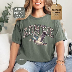 May include: Moss green Auntie t-shirt with a duck graphic. The word "AUNTIE" is printed in a camouflage pattern. The shirt has a relaxed fit and is made of a comfortable material. The shirt is a Comfort Colors 1717.