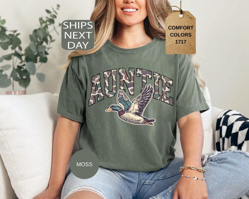 May include: Moss green Auntie t-shirt with a duck graphic. The word "AUNTIE" is printed in a camouflage pattern. The shirt has a relaxed fit and is made of a comfortable material. The shirt is a Comfort Colors 1717.