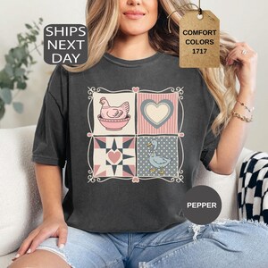May include: Dark grey t-shirt with a patchwork design featuring a hen, heart, and duck. The shirt has the text "COMFORT COLORS 1717" on a tag and "PEPPER" on a circle. The shirt is worn by a person.