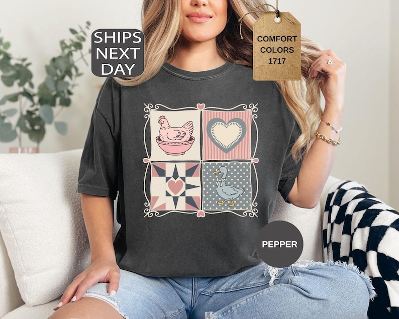 May include: Dark grey t-shirt with a patchwork design featuring a hen, heart, and duck. The shirt has the text "COMFORT COLORS 1717" on a tag and "PEPPER" on a circle. The shirt is worn by a person.