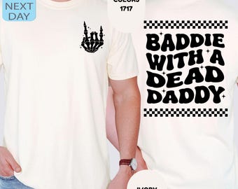 Könnte beinhalten: Ein elfenbeinfarbenes T-Shirt mit einer schwarzen Skeletthand, die auf der linken Brust ein Rock-Geste macht. Die Rückseite zeigt den Text BADDIE WITH A DEAD DADDY in einer wellenförmigen schwarzen Schrift, eingerahmt von zwei schwarz-weißen Karomustern.