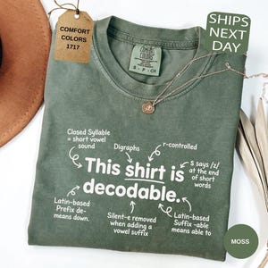May include: A moss green crew neck t-shirt featuring white text that reads, This shirt is decodable, surrounded by linguistic annotations including Closed Syllable, Digraphs, r-controlled, Latin-based Prefix, Silent-e, and Latin-based Suffix.