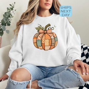 May include: White sweatshirt featuring a colorful patchwork pumpkin design with a bow and stem. The pumpkin is made of various patterned fabrics in orange, green, and brown. The text "SHIPS NEXT DAY" is visible.